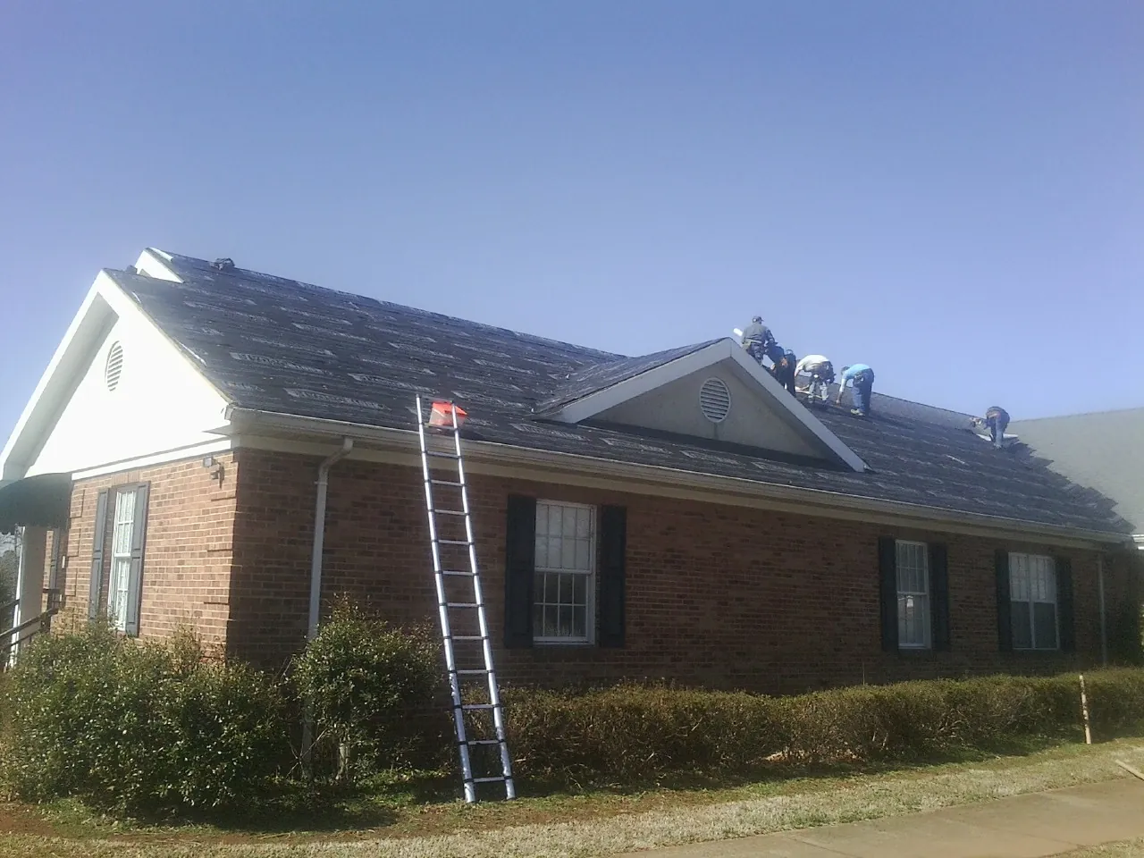 Crew tearing shingles off a brick ranch roof with a ladder set — Emergency Roof Repair work by our crew in Discovery Bay