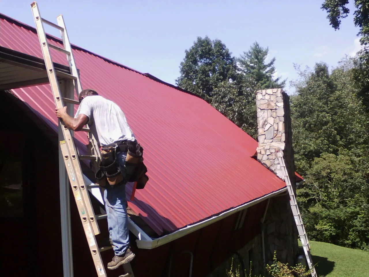 Roofer on a ladder beside a red metal roof and stone chimney — Soffit & Fascia Repair by Roofing Brothers in Discovery Bay